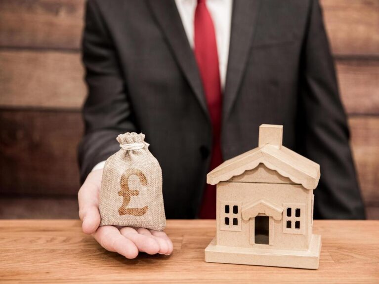 Concept showing cost house value increase with solar panel installation. Man holding small bag with pound sign next to small model of house. Blog image for More Than Solar Panels
